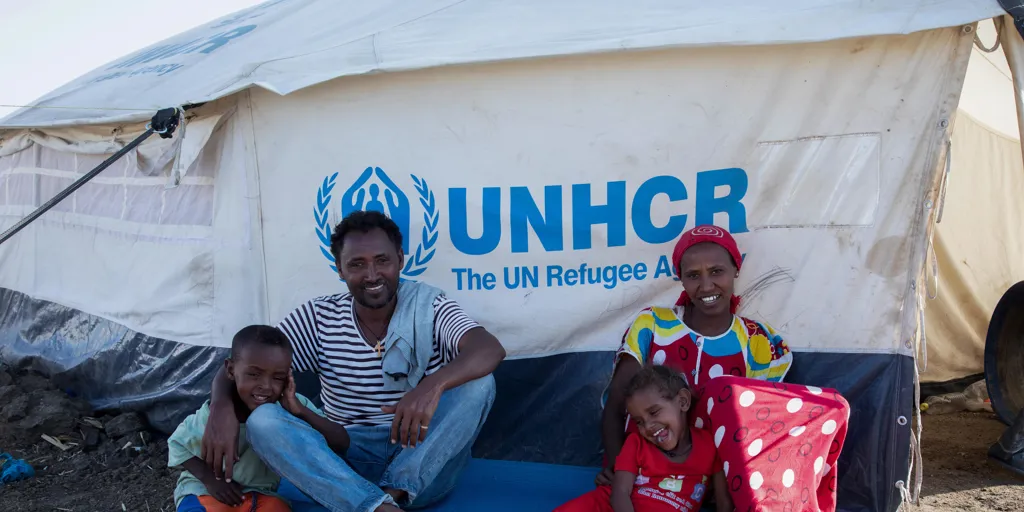 Family of Ethiopian refugees smiling. UNHCR tent in the background. Ethiopian refugees Tsiruy Dagnew, 40, Abeba Kalayu, 32, with their children Semere, 5, and Eleni, 3, enjoy the sun near their tent at Tunaydbah camp in Sudan. Many of its refugees have found innovative and resourceful ways to begin rebuilding their lives.