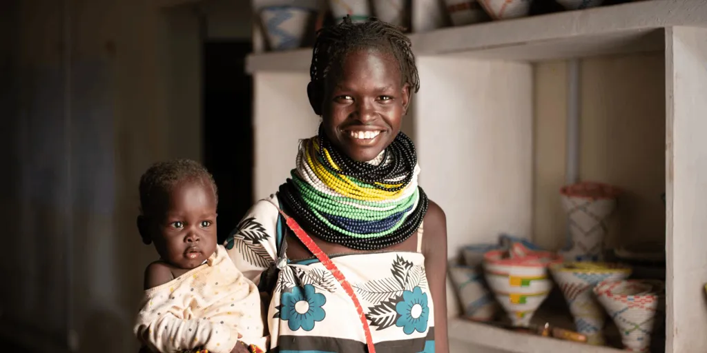 Lucy Akai, a Kenya woman, holds her baby and smiles at the camera. She is wearing brightly coloured beaded jewelery.